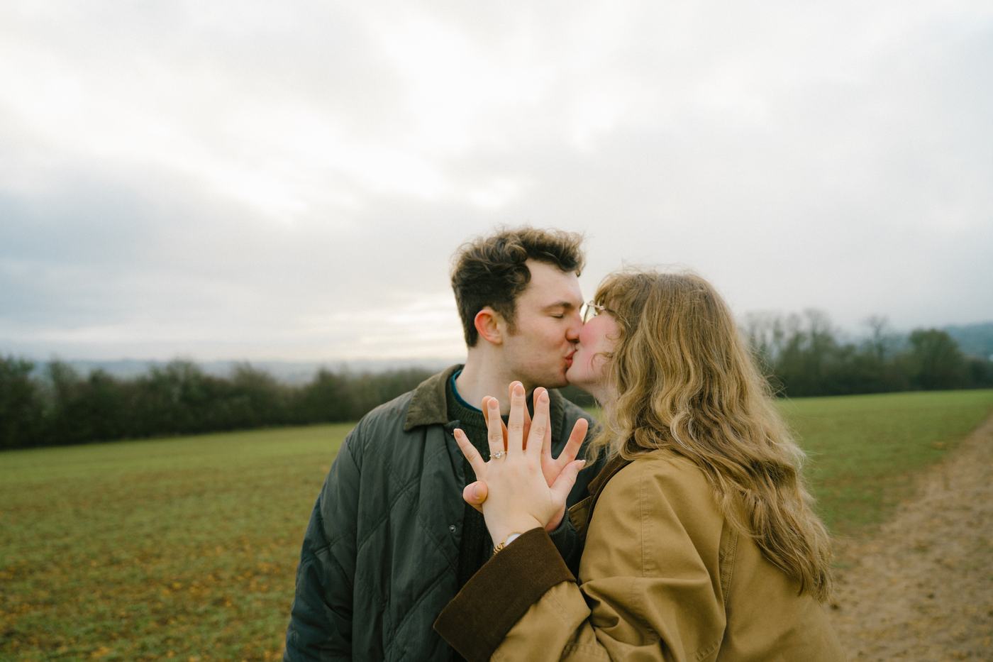 Couple kissing in a green field, showing off an engagement ring