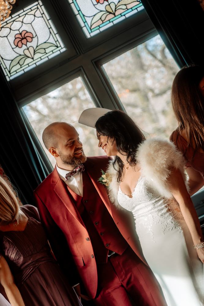 Groom in a red suit embracing the bride by a stained-glass window